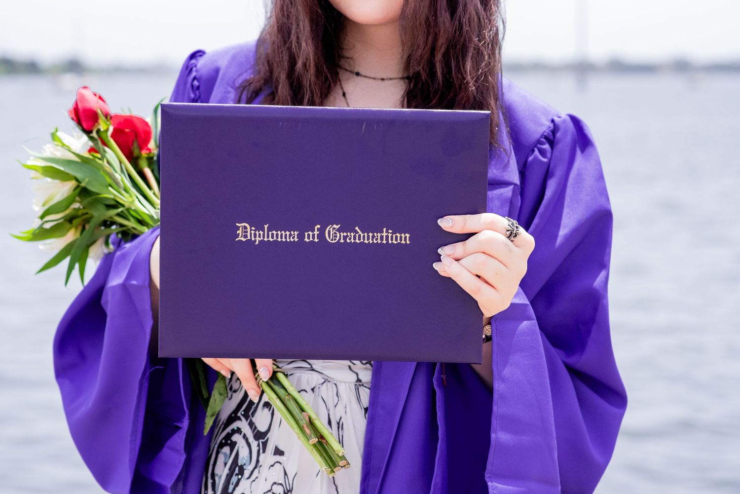 Graduate in purple gown holds Diploma of Graduation and red roses outdoors near water.