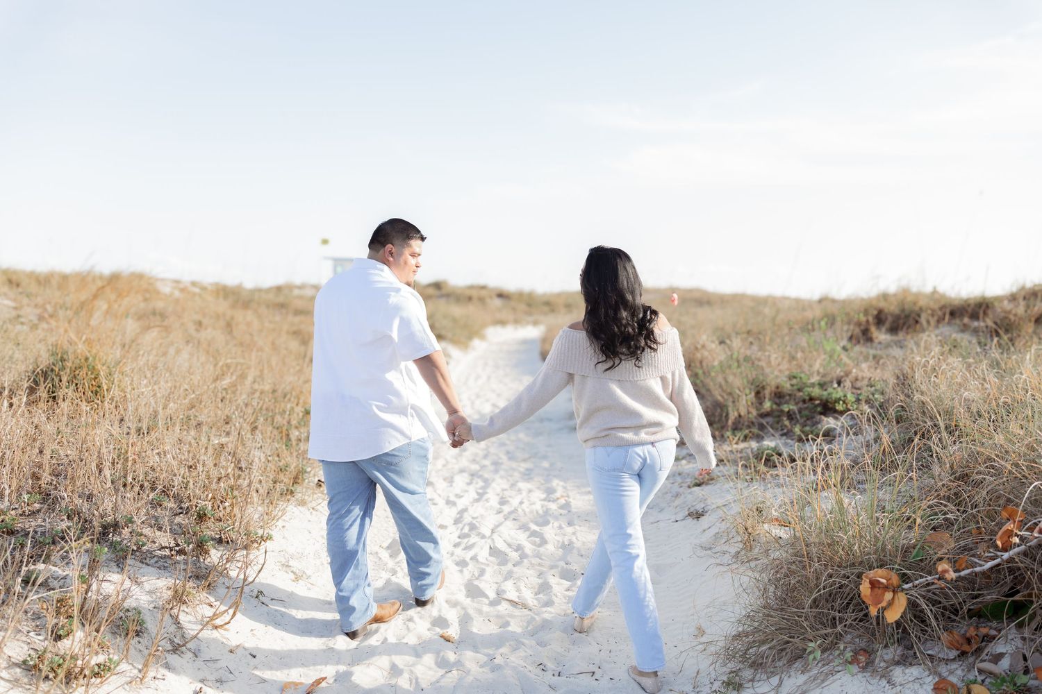 Couple holding hands walking on a sandy beach path surrounded by dunes and sea grass on a sunny day.