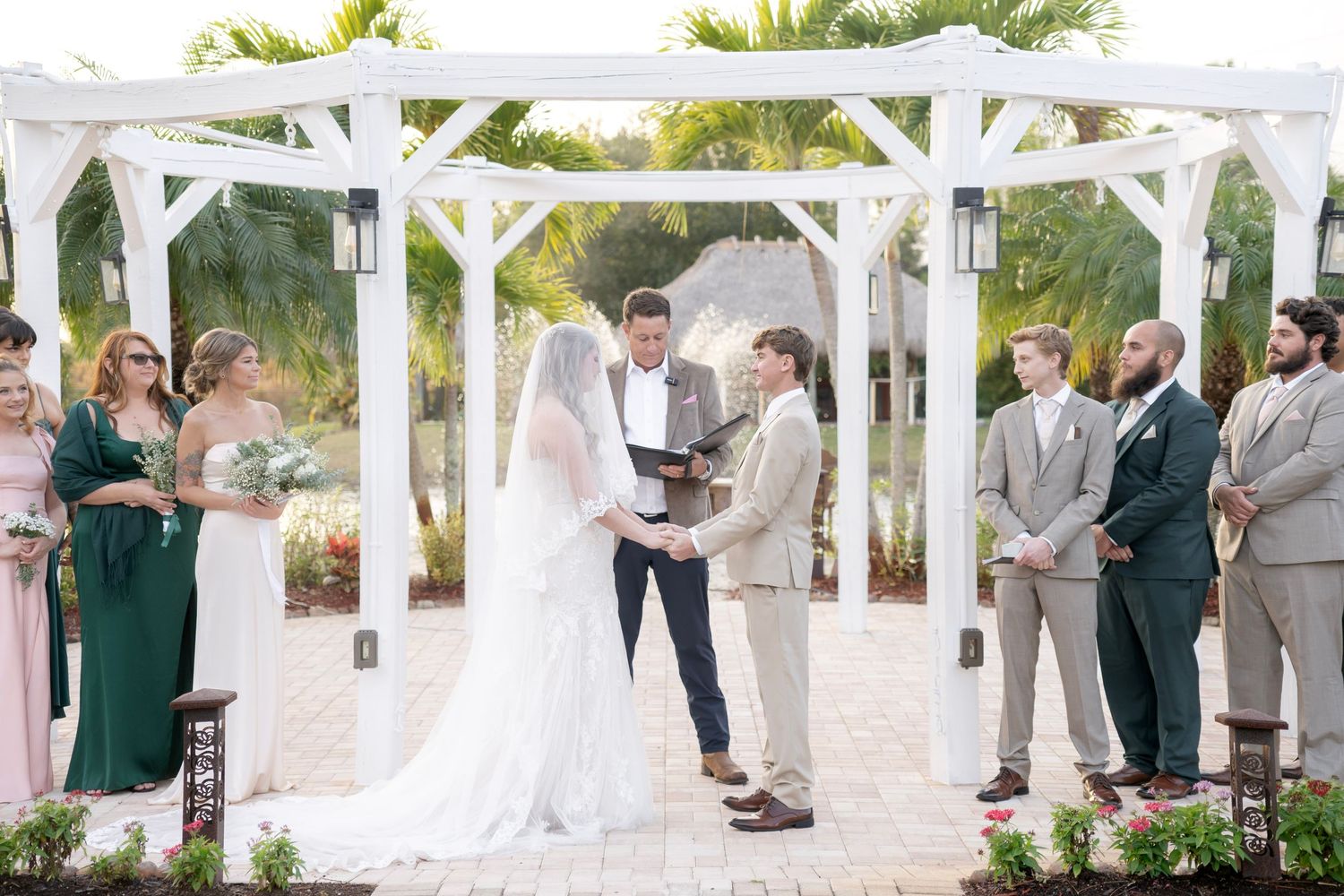Outdoor wedding ceremony under white pergola with bride, groom, officiant, and wedding party.