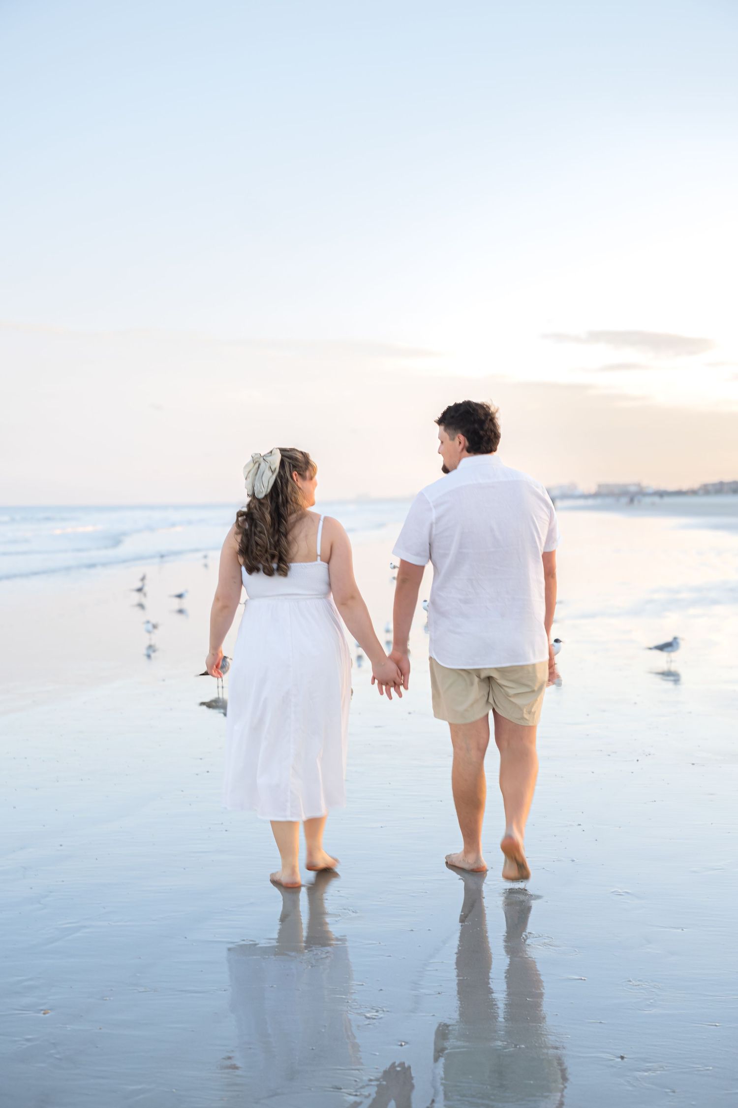 Couple holding hands walking barefoot on a serene beach at sunset, woman in white dress, man in white shirt.