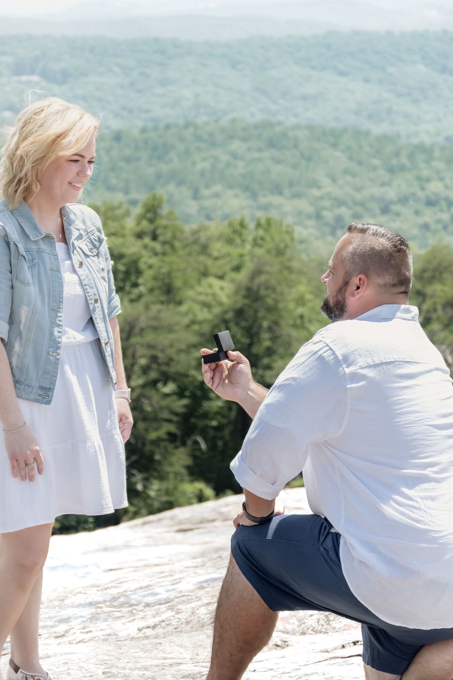Man kneeling on rocky overlook proposing with ring box to smiling blonde woman with forest mountains backdrop.