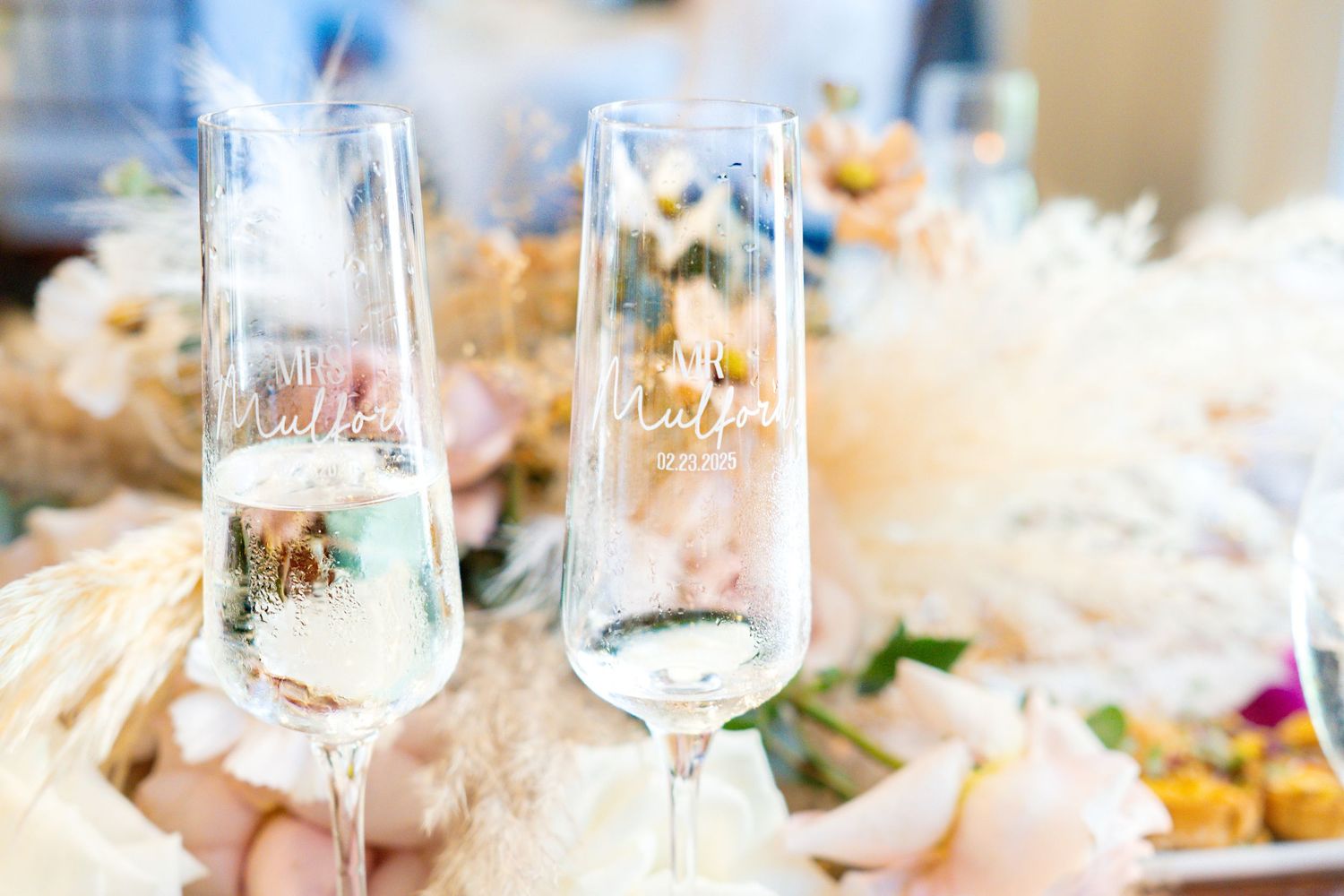 Two elegant personalized champagne flutes with gold text at a floral wedding reception table.