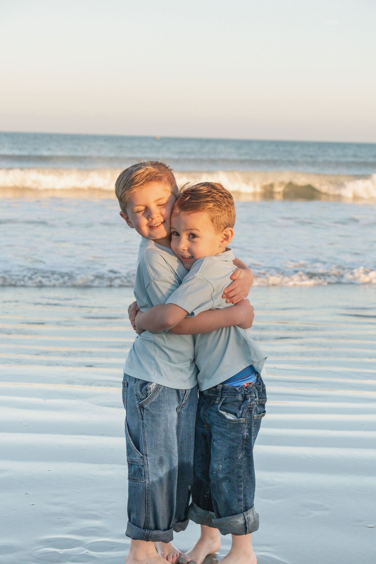 Two young boys hugging each other on a sandy beach with ocean waves in the background at sunset.