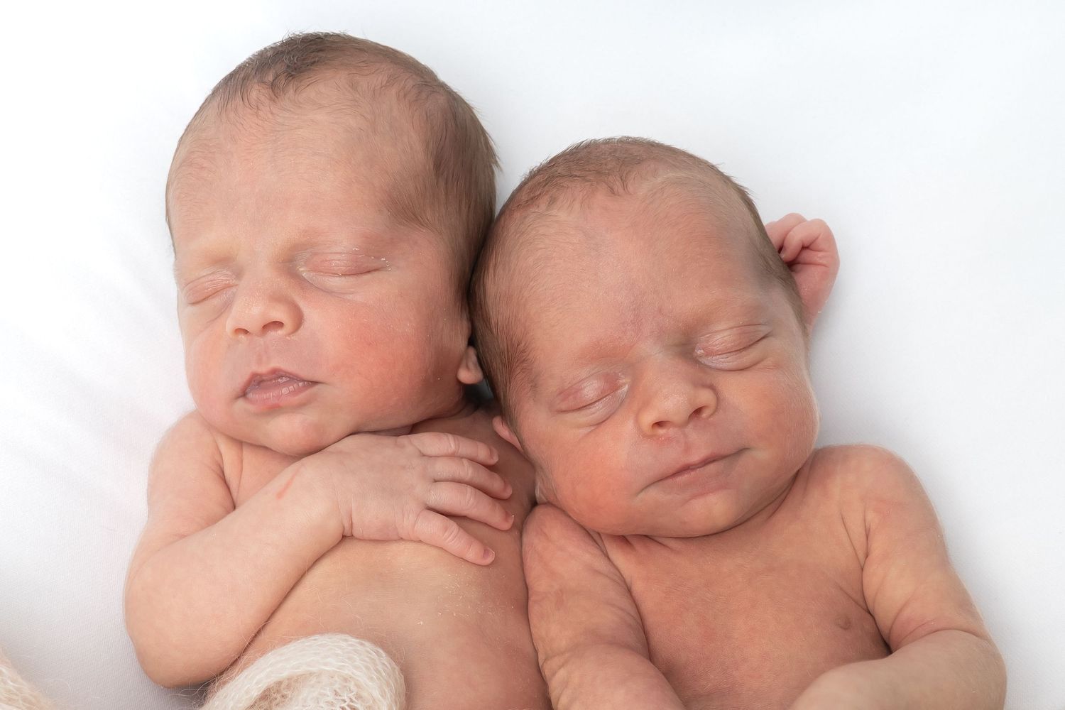 Two sleeping newborn twins lying side by side on a white blanket in a peaceful newborn photography pose.