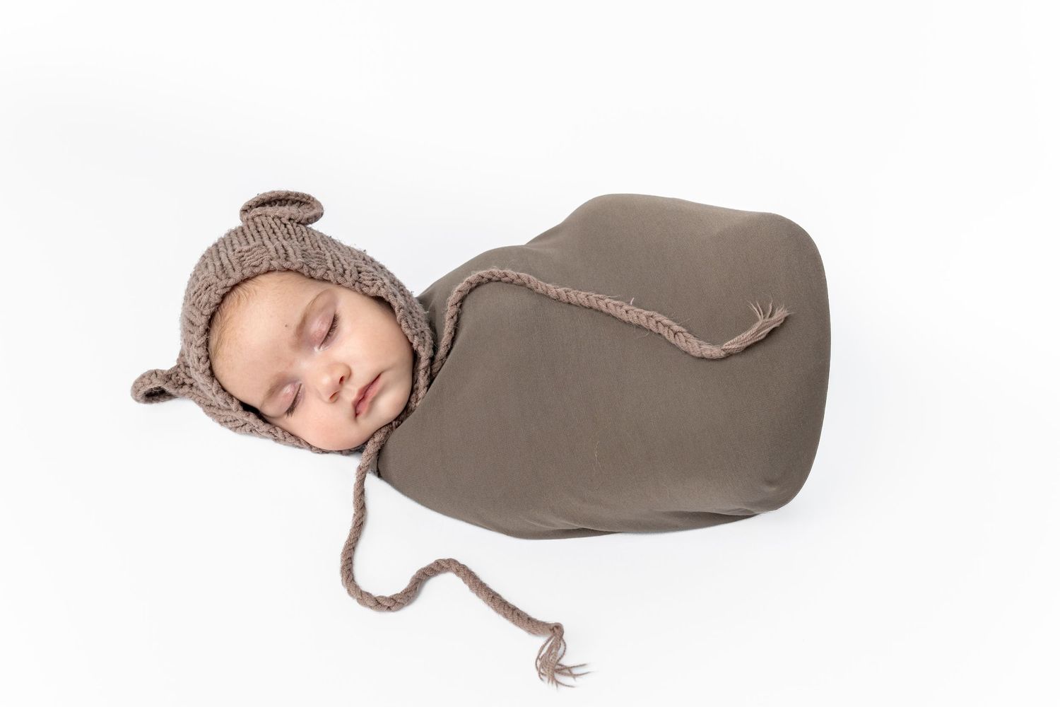 Sleeping newborn baby wrapped in olive green swaddle wearing a knitted brown bear bonnet on white background.