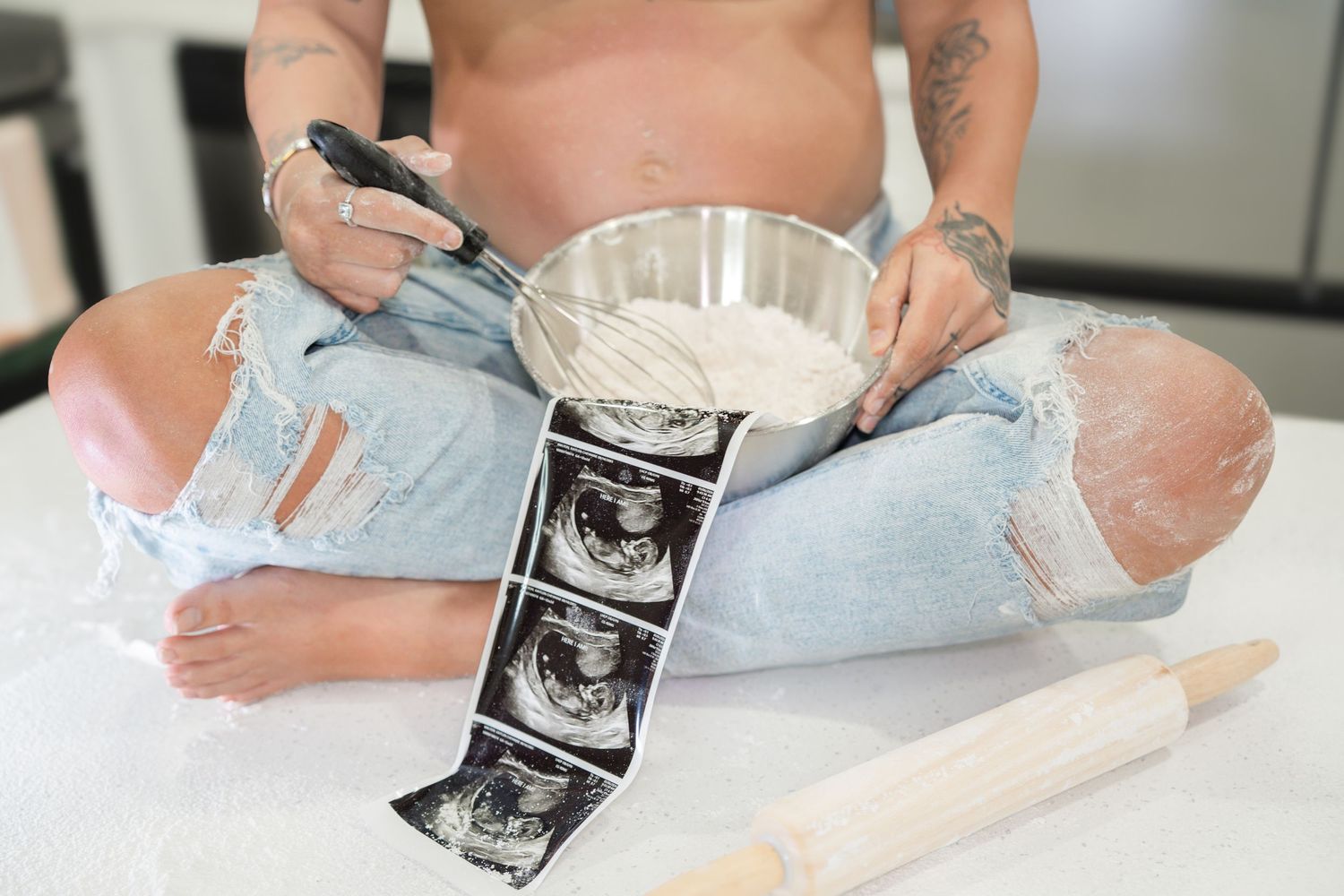Pregnant person sitting cross-legged with ultrasound photos, mixing bowl, and rolling pin on white surface.