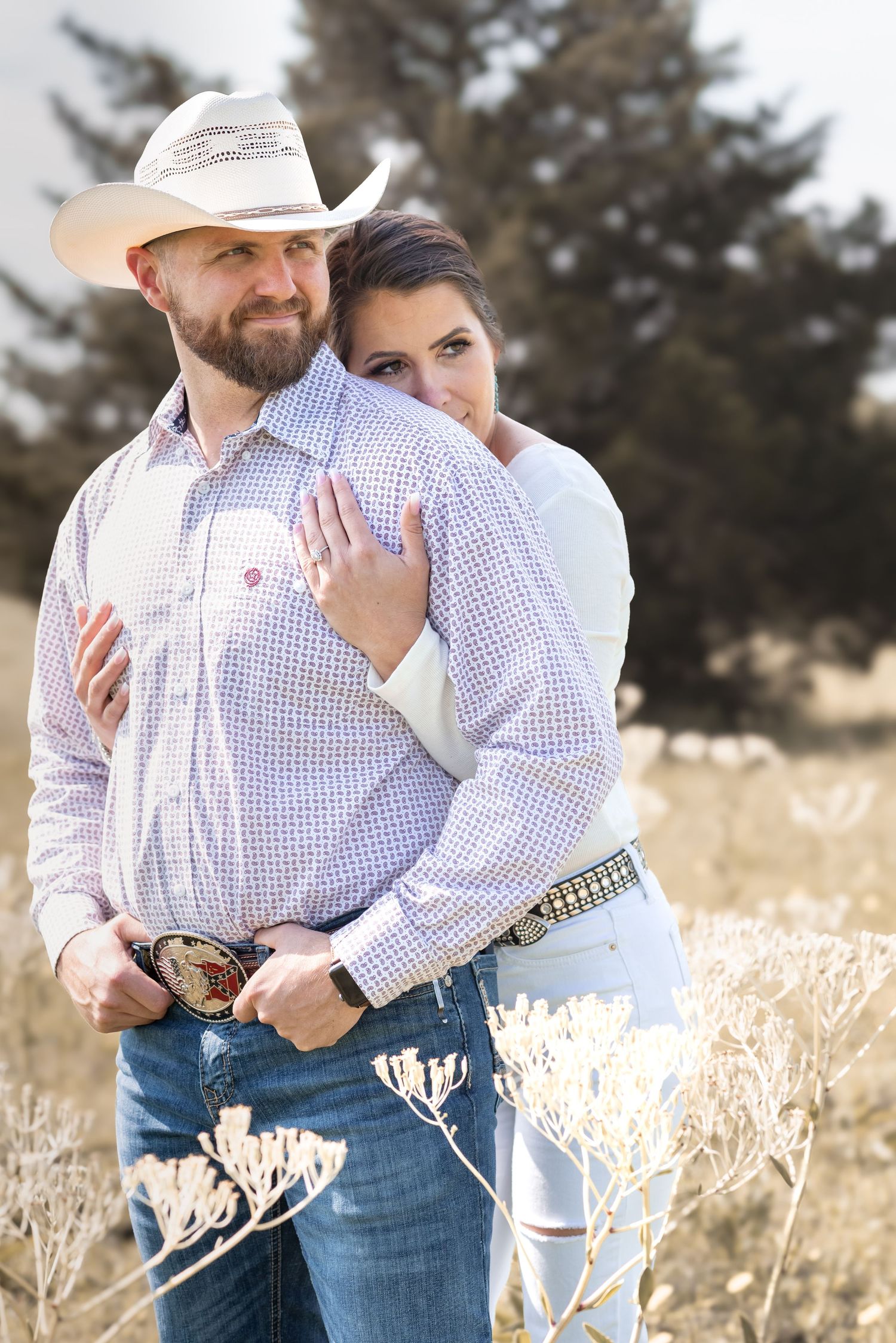 Couple embracing in a field, man wearing cowboy hat and checkered shirt with decorative belt buckle.