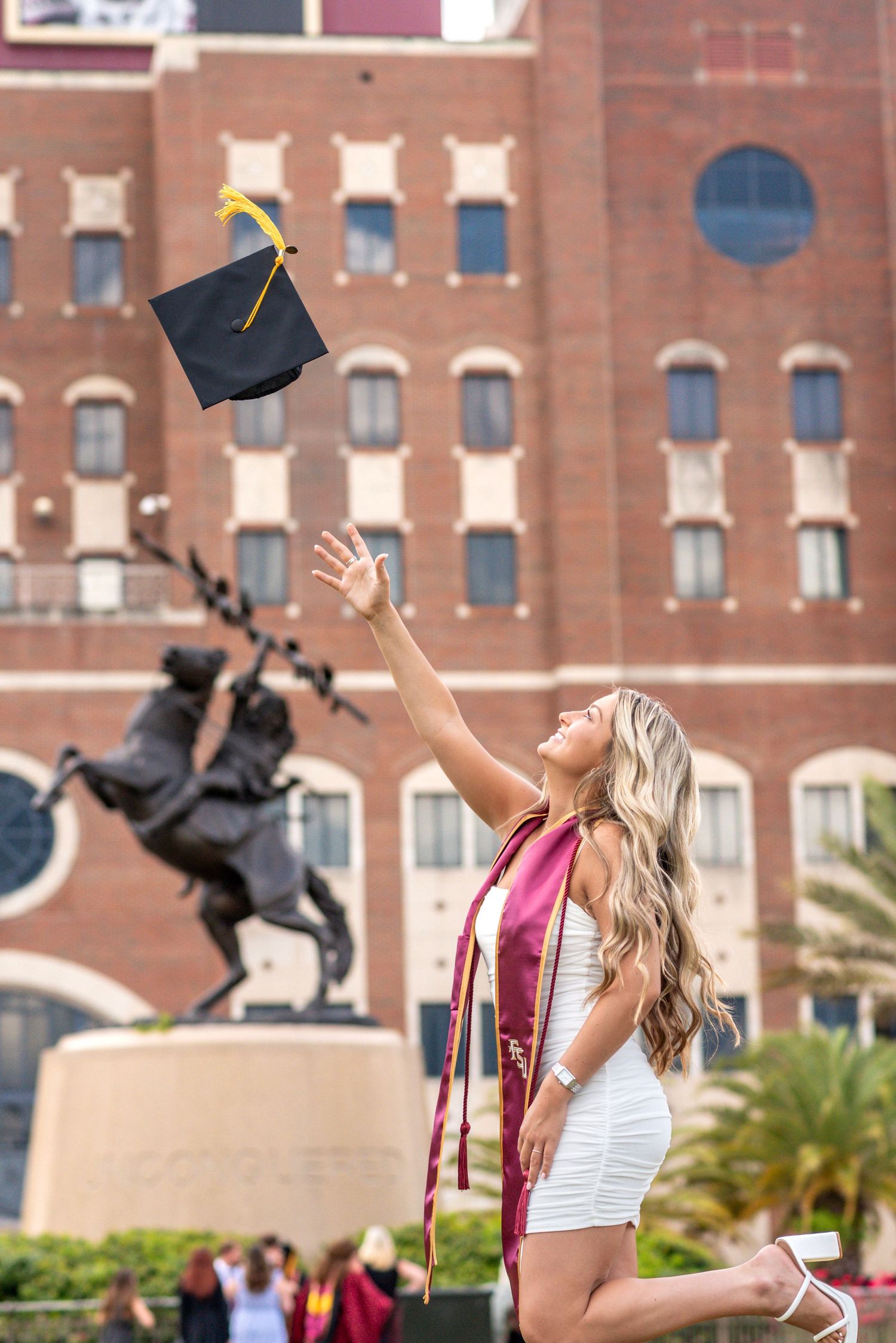 Graduate in white dress tosses cap near Unconquered statue at Florida State University on graduation day.
