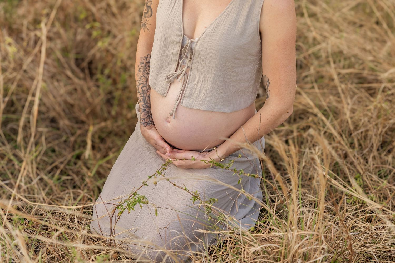 Pregnant woman in beige dress kneeling in golden grass field, cradling baby bump with both hands.