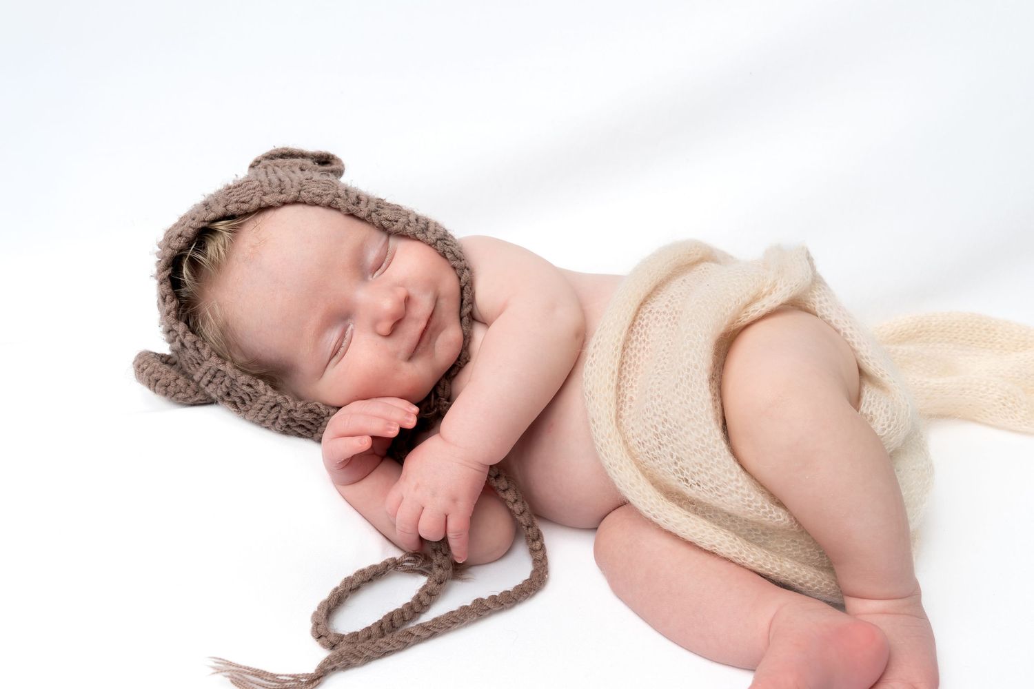 Sleeping newborn baby wearing a cute knitted bear ear hat, wrapped in a soft beige blanket on white background.