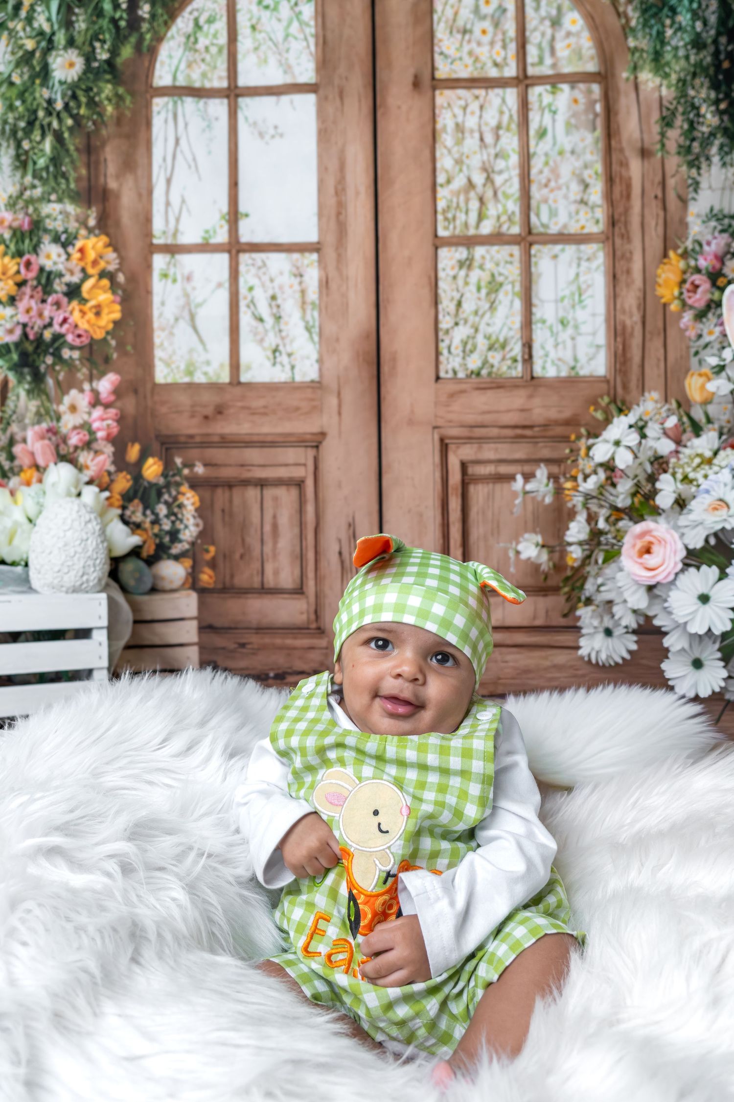 Smiling baby in green gingham outfit and hat sits on white fur blanket with floral wooden door backdrop.