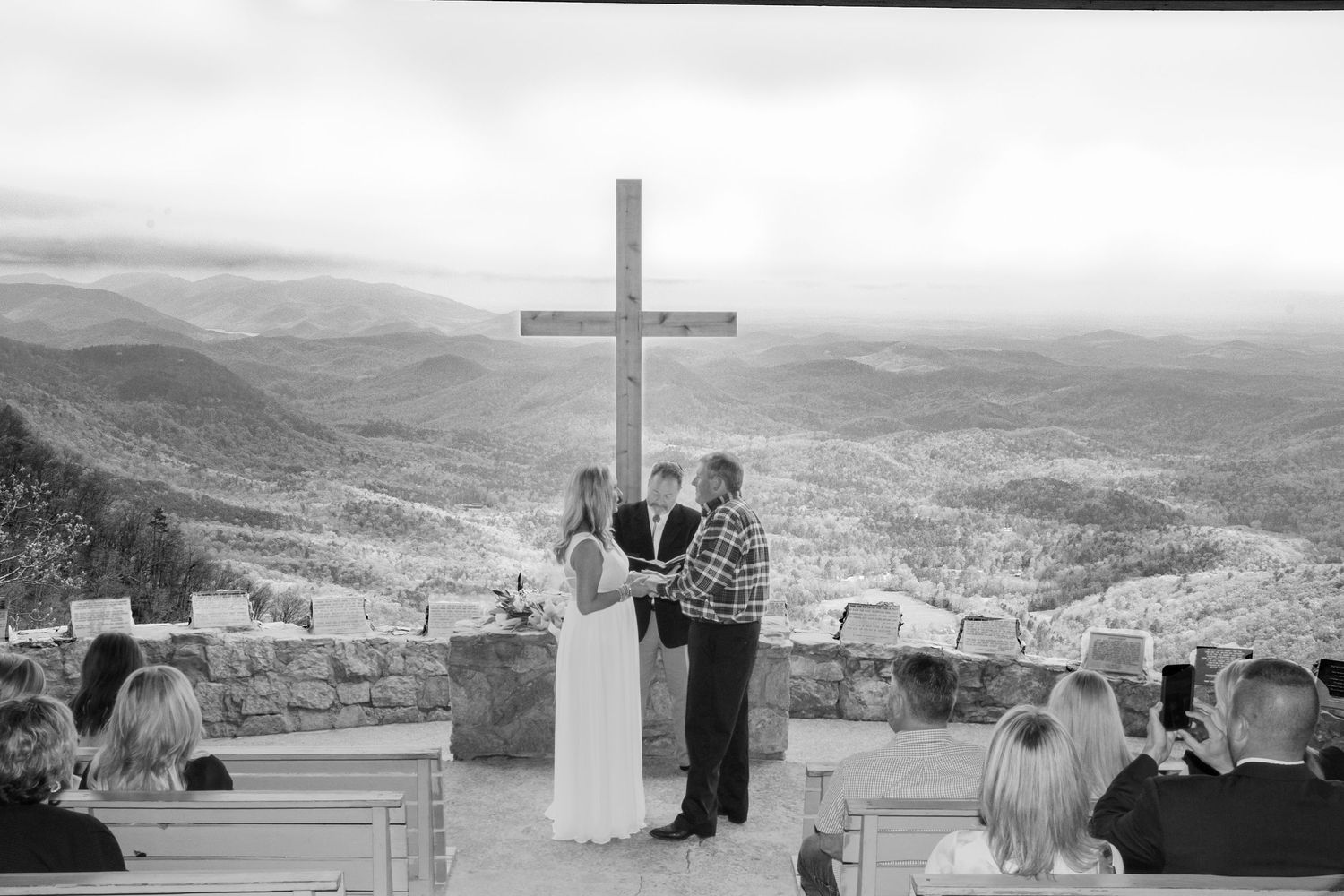 Black and white outdoor wedding ceremony with couple at stone altar, large cross, and stunning mountain valley views.