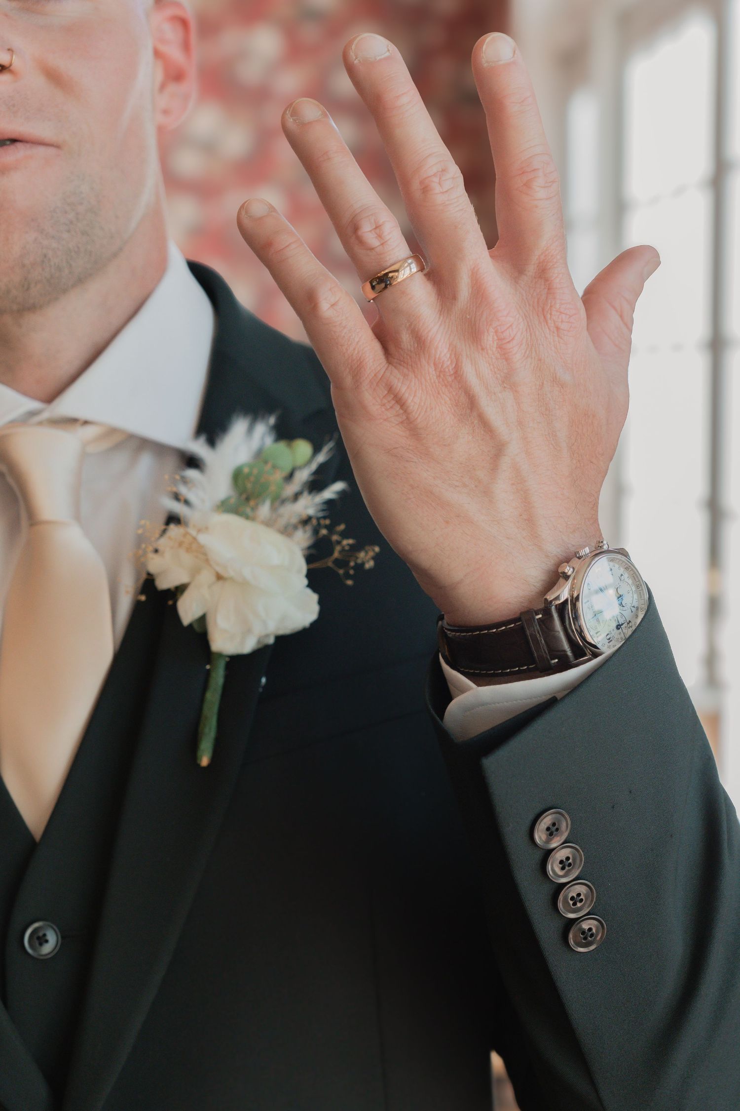 Groom in dark suit displaying wedding ring on hand with boutonniere and luxury watch visible.