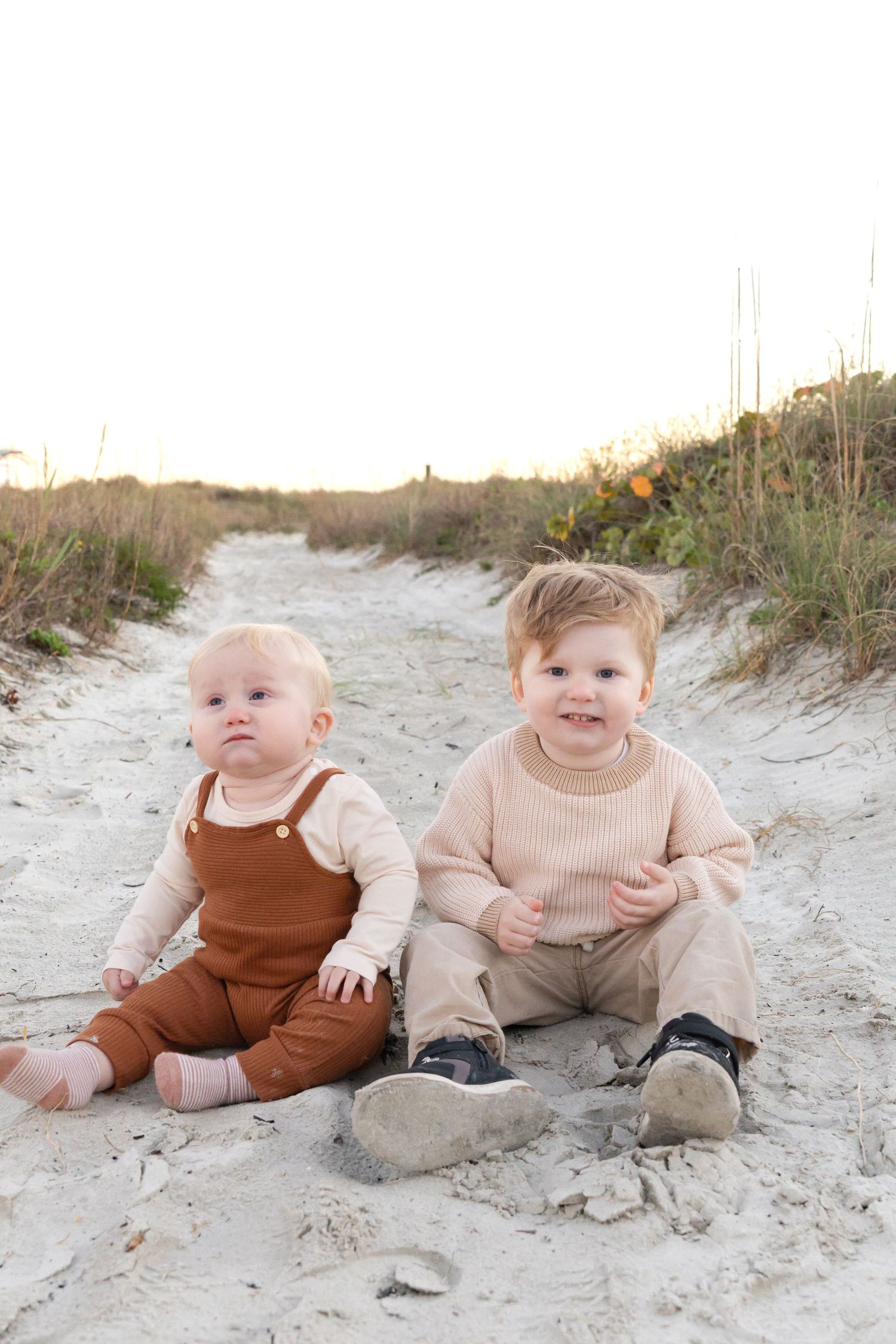 Two young toddlers sitting on sandy beach path, wearing warm earth-tone outfits, with dune grass in background.