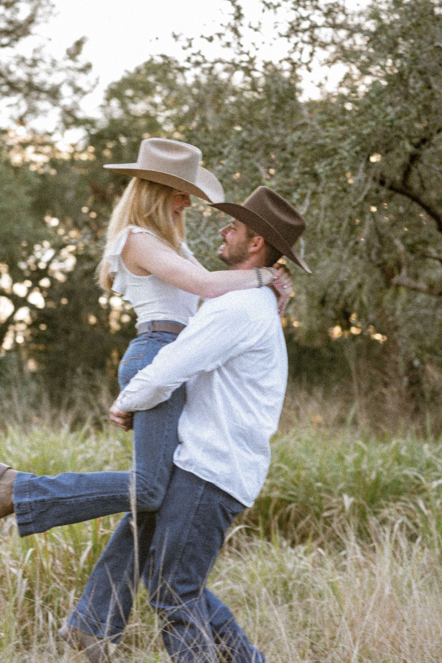 Couple in cowboy hats embrace in a golden field, man lifting woman as they share a romantic moment outdoors.