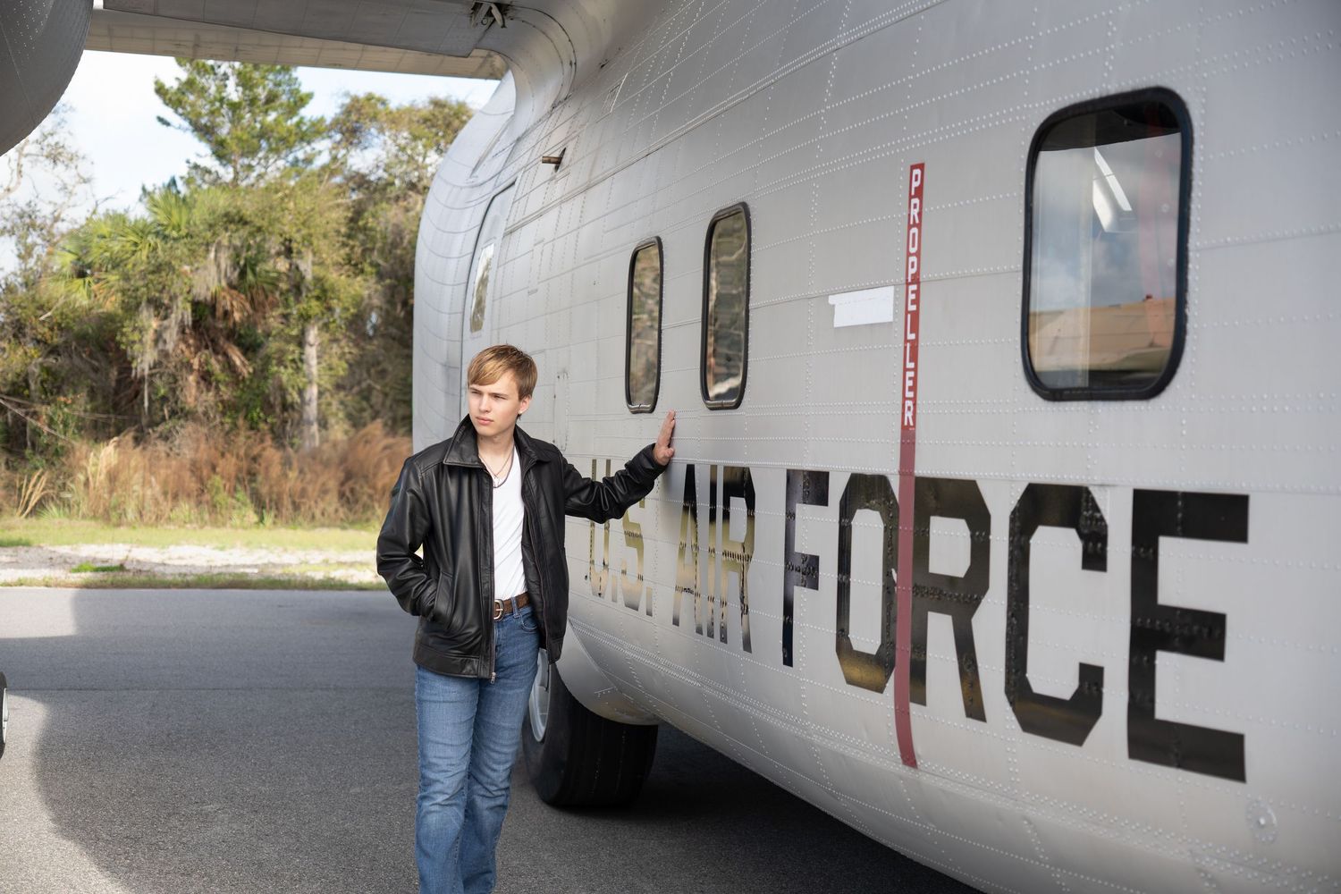 Young man in leather jacket posing beside US Air Force military aircraft outdoors at airfield.