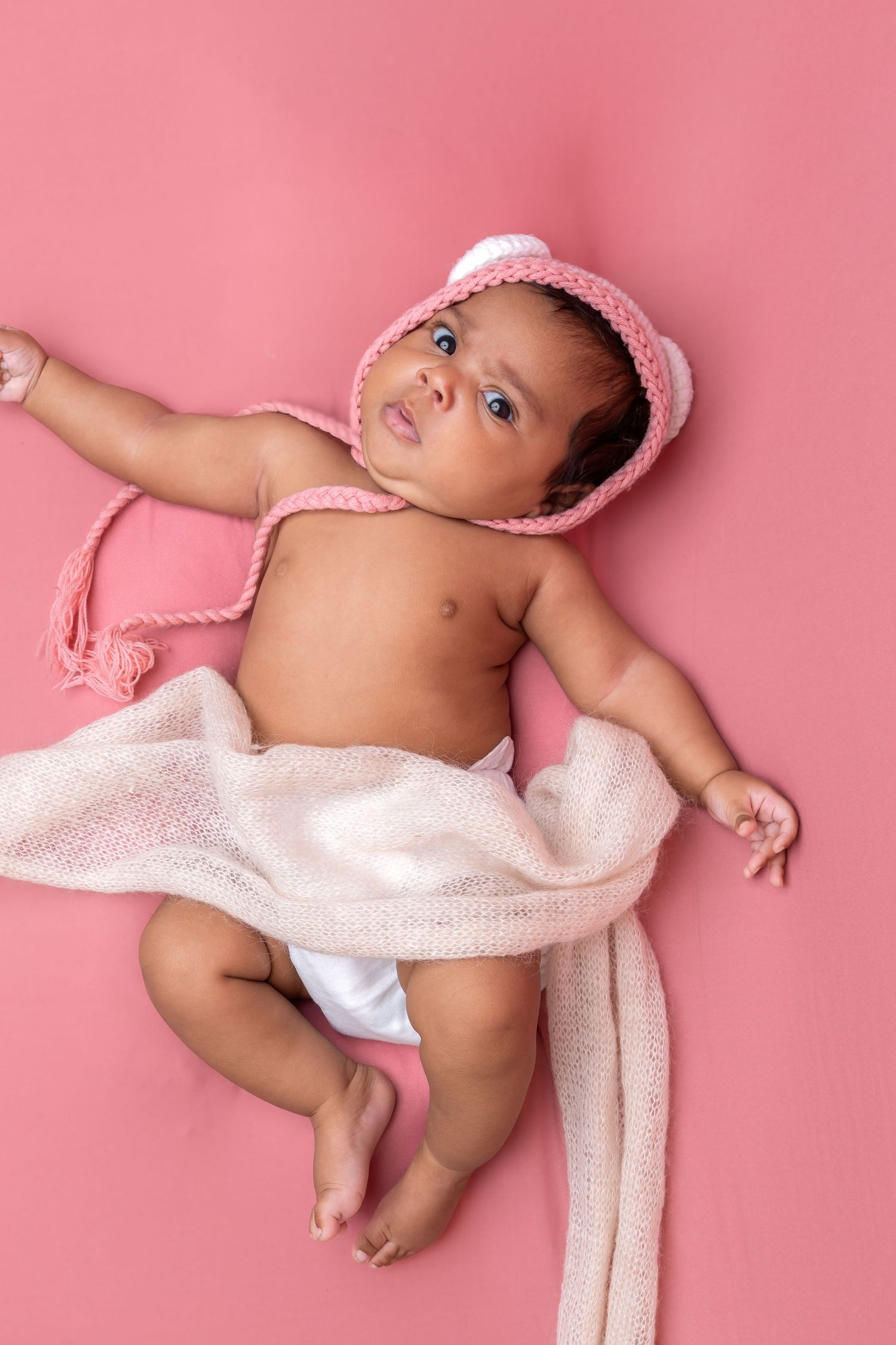 Newborn baby girl in pink knit bonnet lying on pink background wrapped in white gauze blanket.