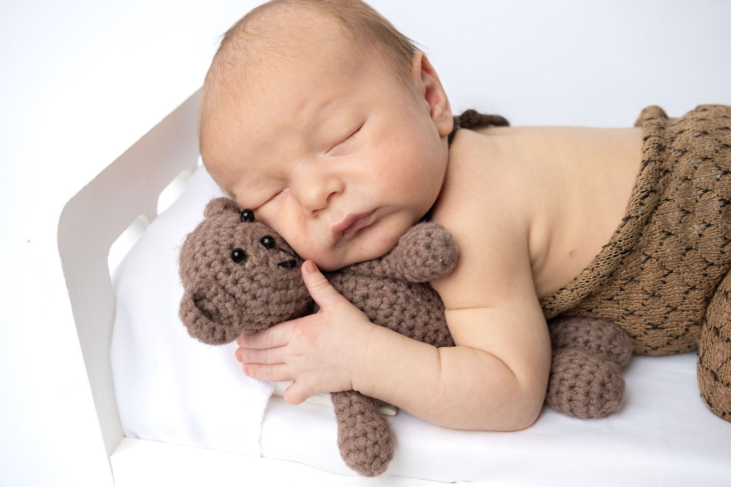 Sleeping newborn baby cuddling a brown crocheted teddy bear toy, wrapped in a knit blanket on a white bed.