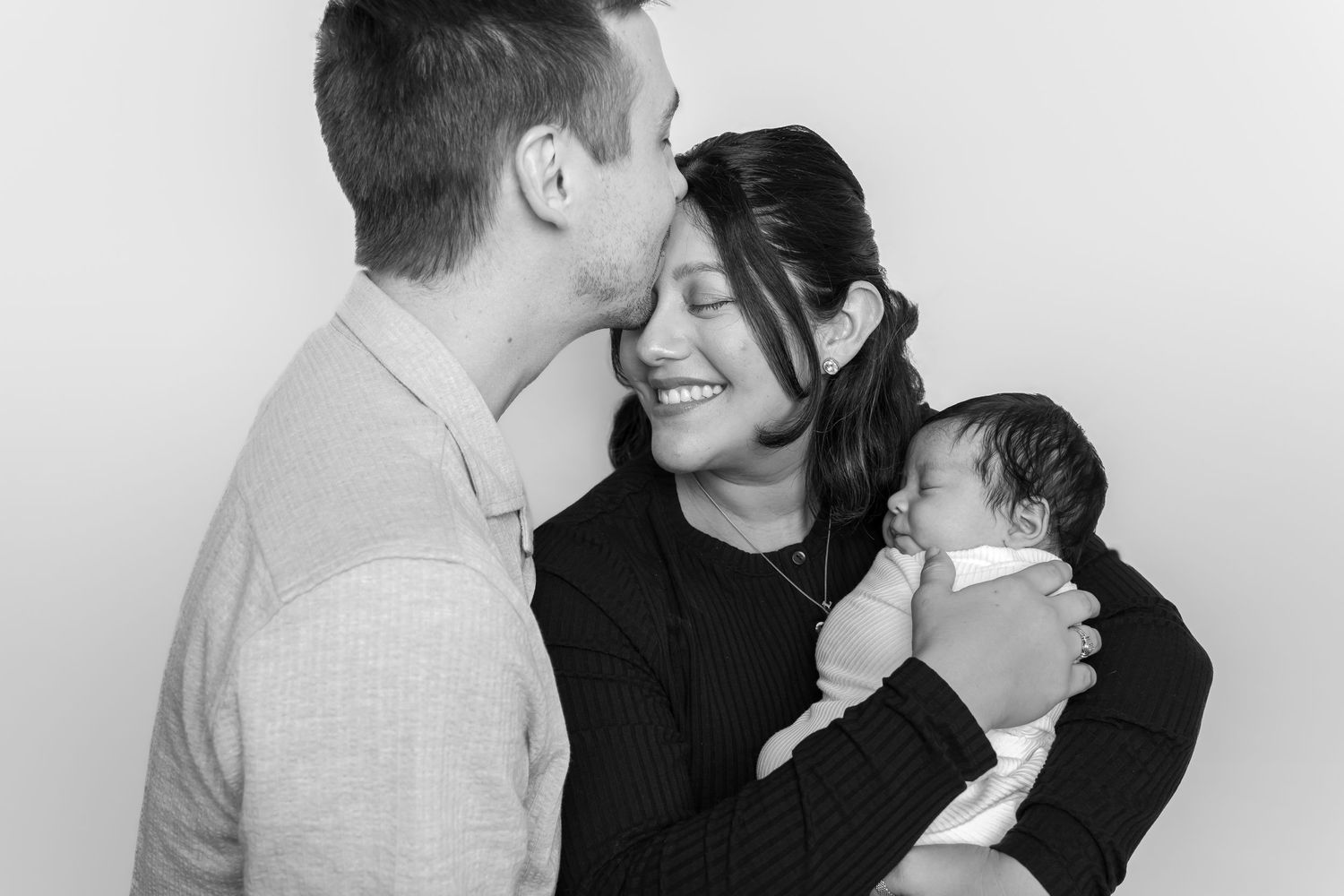 Black and white photo of a couple smiling and kissing their newborn baby in a studio portrait.