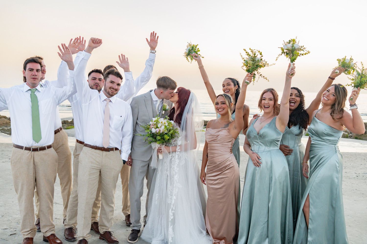 Wedding party celebrating on beach, bride and groom kissing while bridesmaids in sage green dresses and groomsmen cheer.