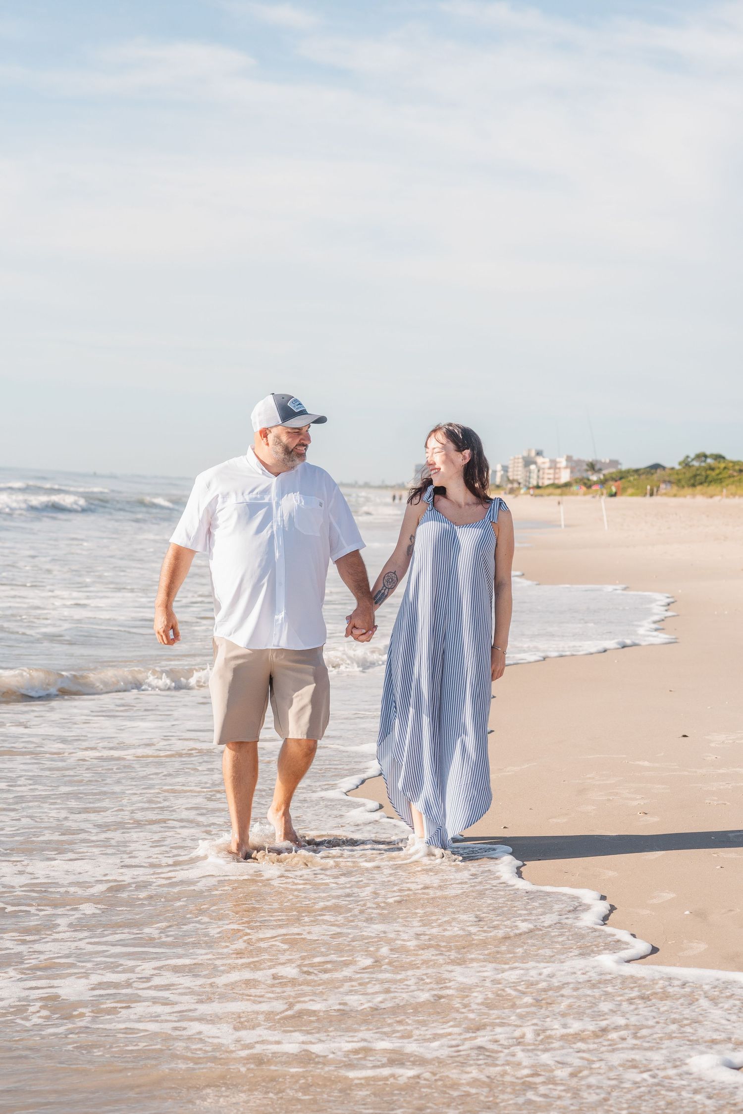 Couple holding hands walking along a sunny beach shoreline, man in white shirt and cap, woman in blue maxi dress.