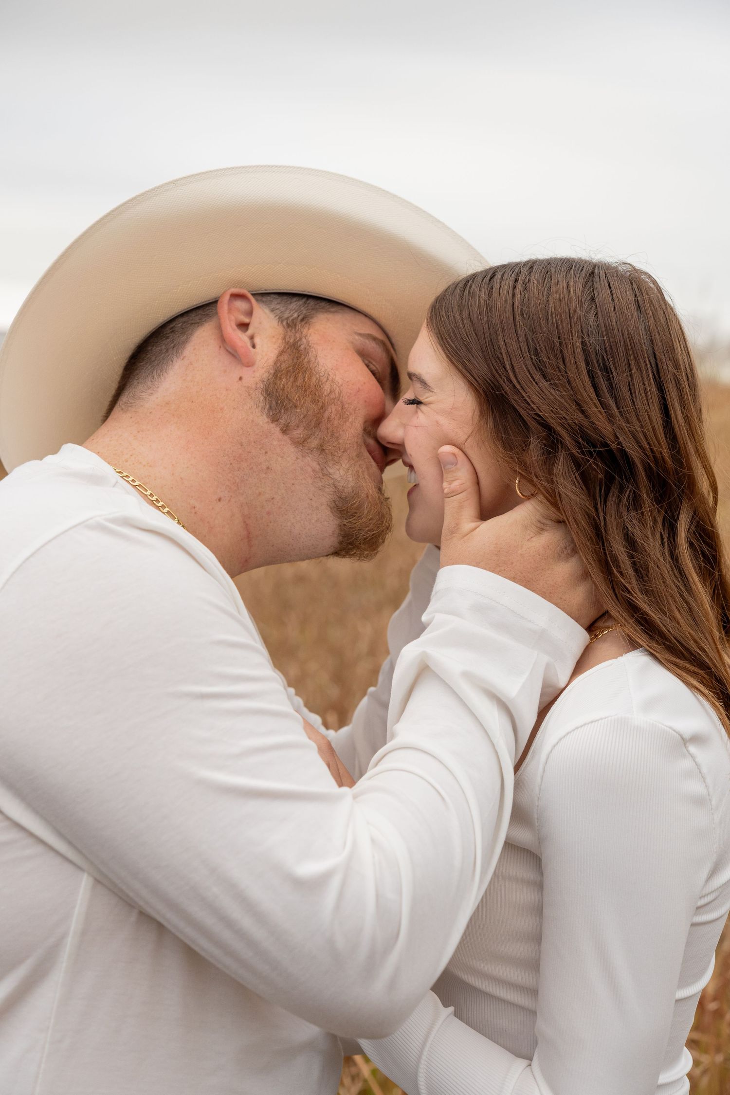 A couple in matching white outfits shares a romantic kiss, the man wearing a cowboy hat and gold chain.