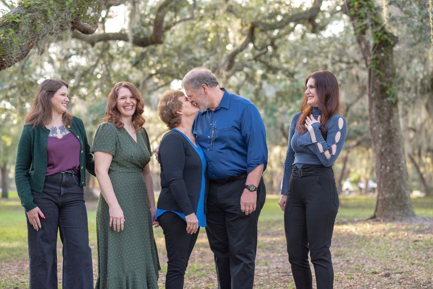 Family of five laughing and sharing a tender moment outdoors under oak trees in a lush park.