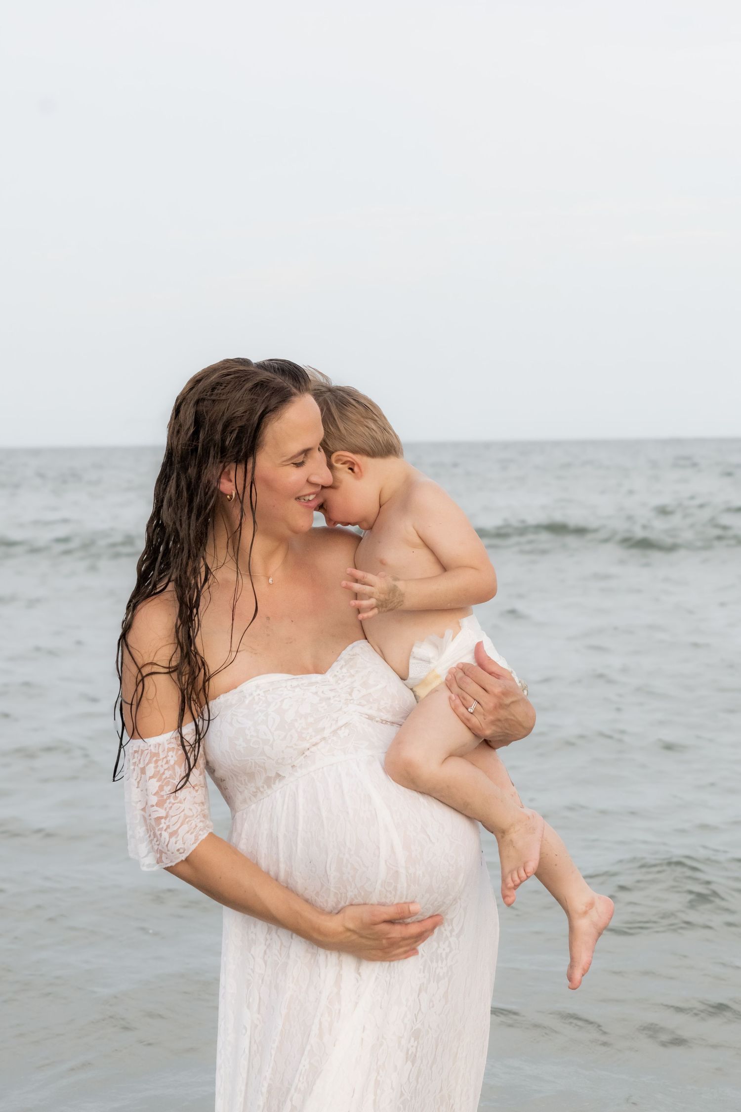 Pregnant woman in white lace dress holds toddler on beach, sharing tender moment with ocean waves behind them.