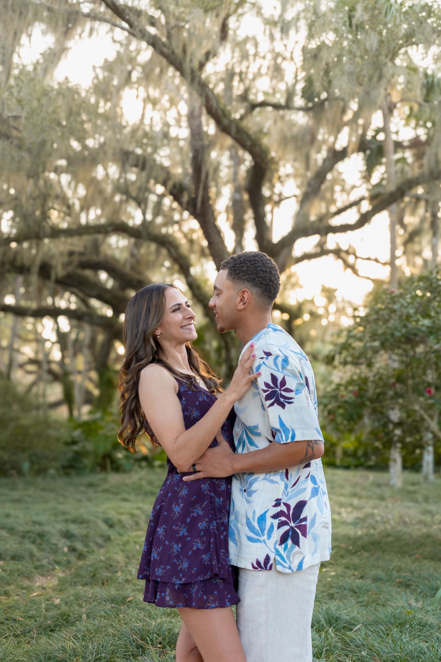 Couple embracing and smiling under a large moss-draped oak tree at golden hour in a lush green park.