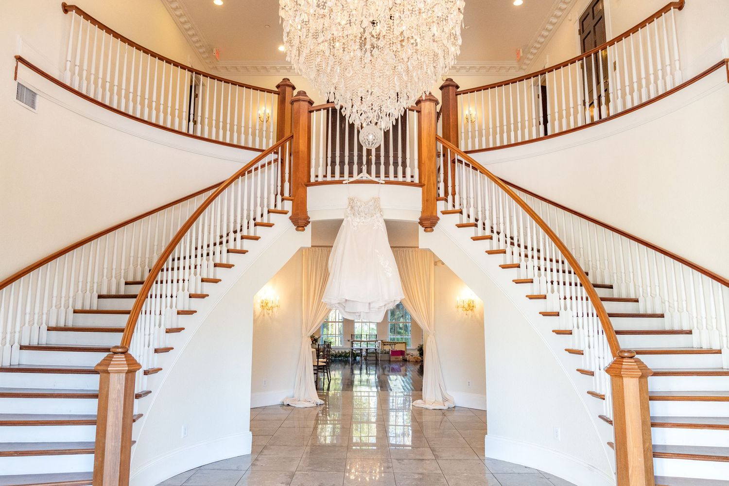 Elegant grand foyer with dual curved staircases, crystal chandelier, and a wedding dress displayed between the stairs.