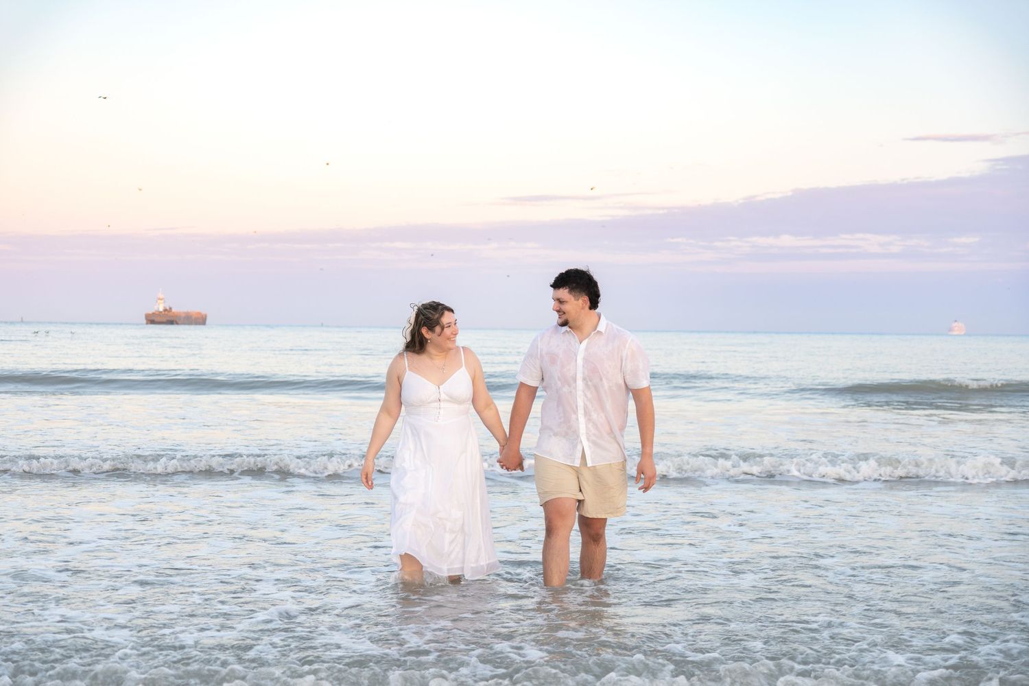 Romantic couple walking hand in hand through shallow beach waves at dusk, dressed in white and khaki attire.