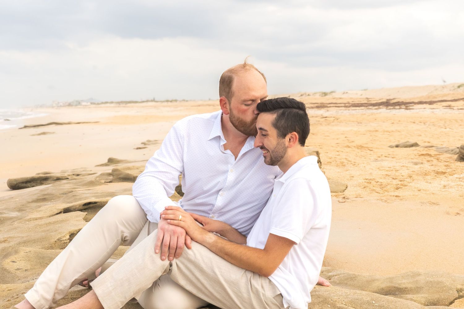 Two men share a tender moment sitting together on rocky beach with sandy shore and cloudy sky backdrop.