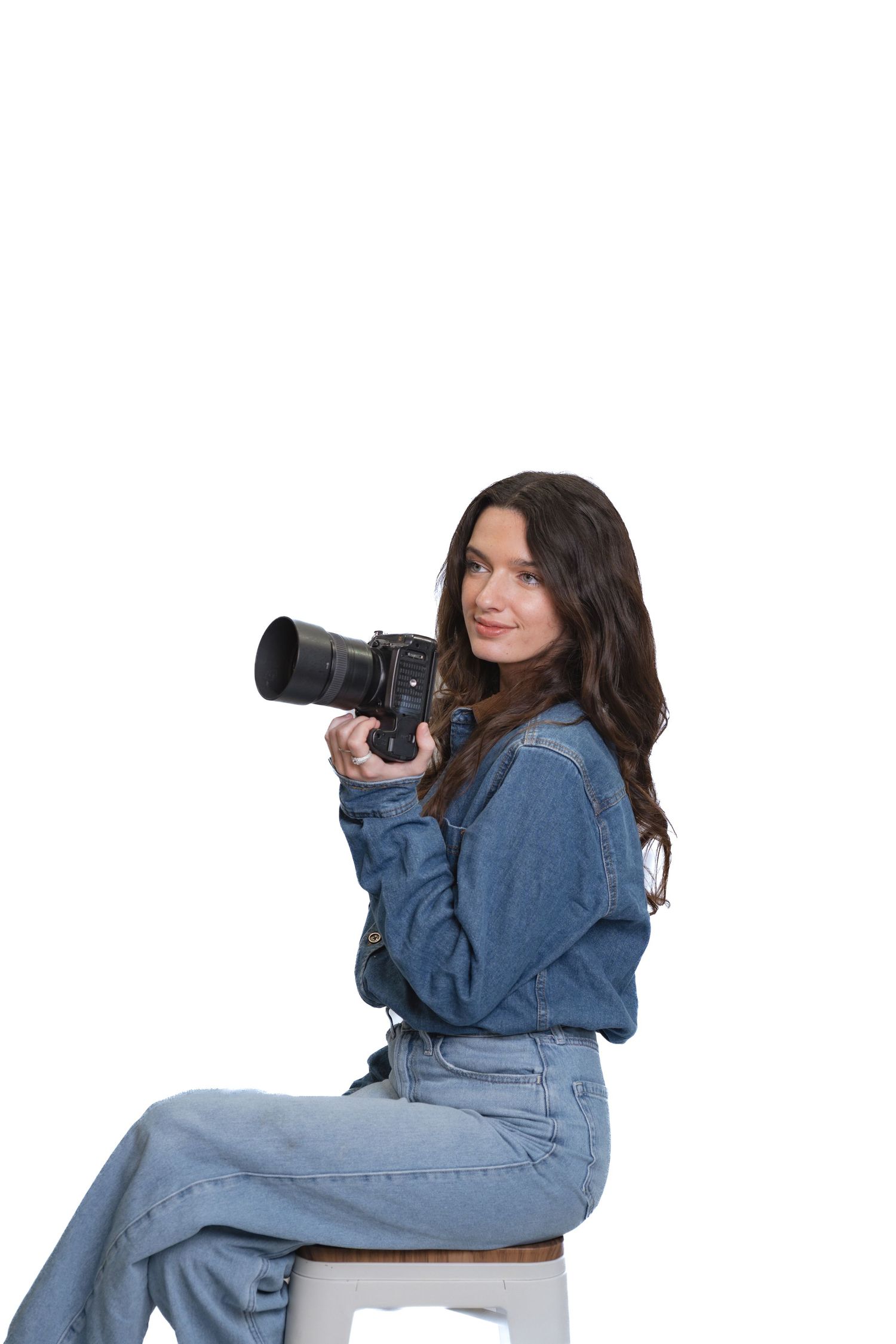 Brunette female photographer in denim outfit seated on stool with DSLR camera on white background.