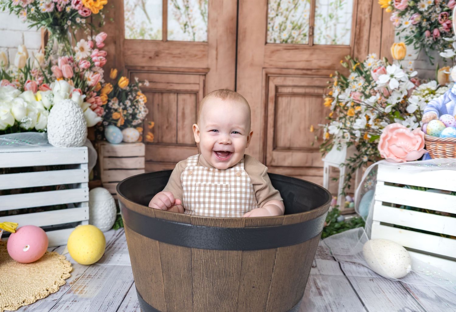 Happy laughing baby sitting in rustic wooden barrel with colorful Easter egg spring decorations.