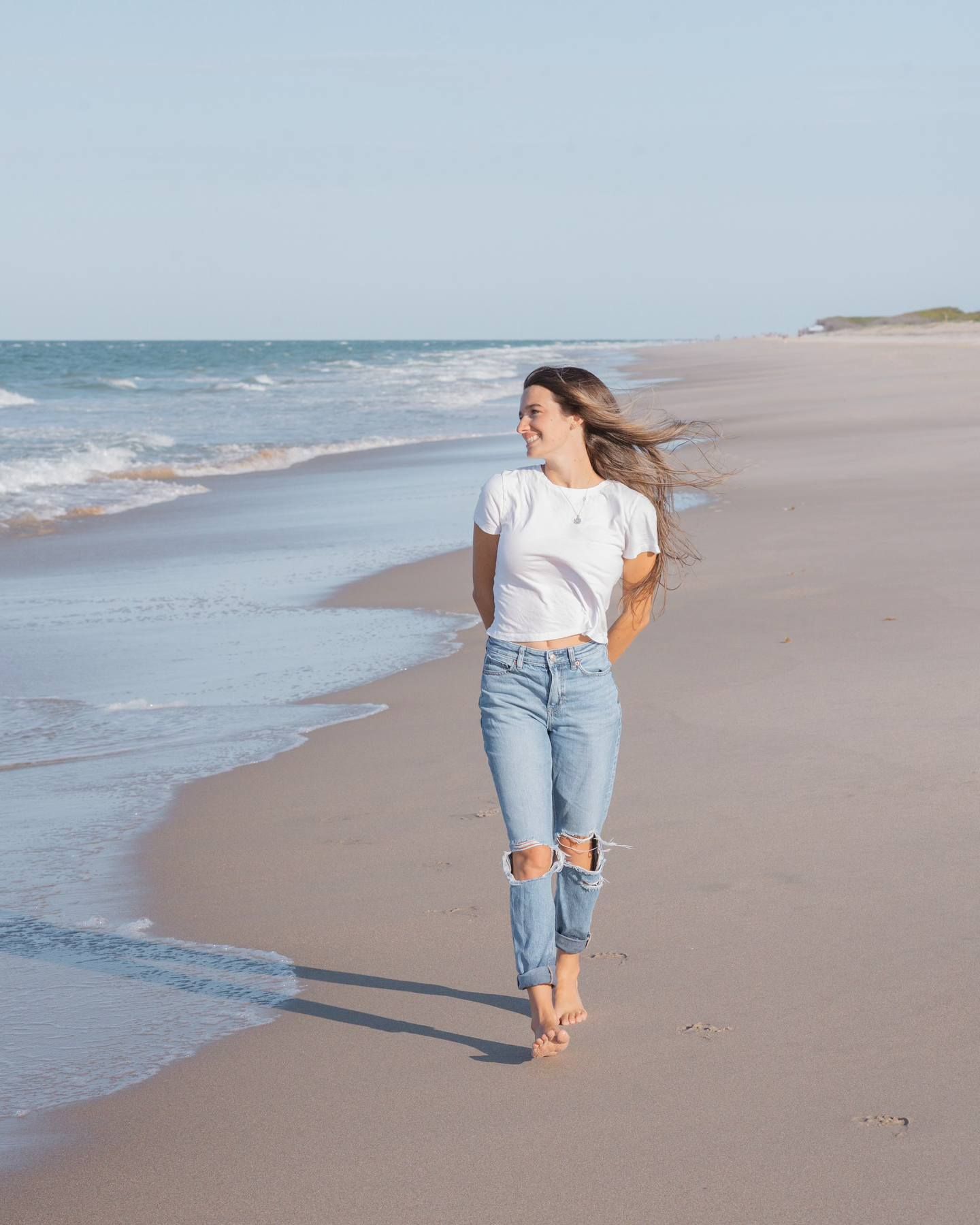 Smiling woman in white t-shirt and ripped jeans walking barefoot along a serene sandy ocean beach.