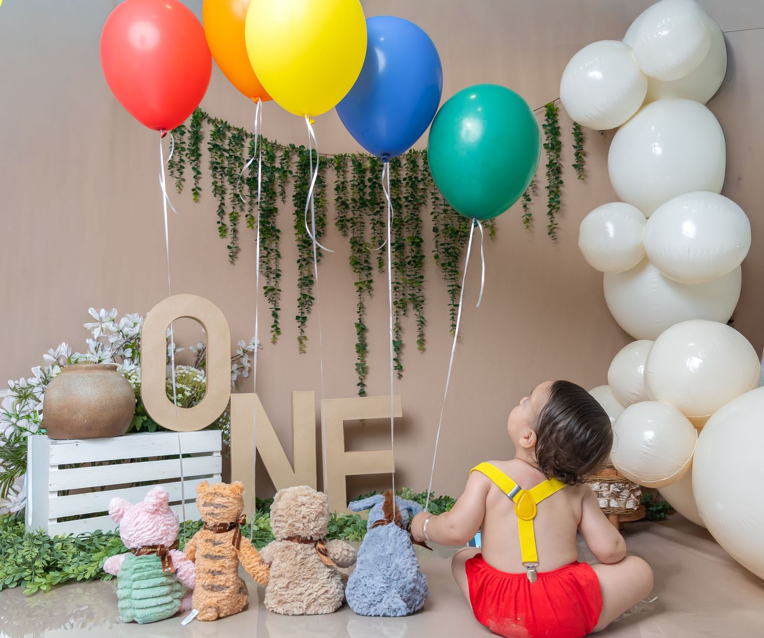 Baby boy in red outfit sitting in front of colorful balloons and ONE letters for a first birthday celebration.