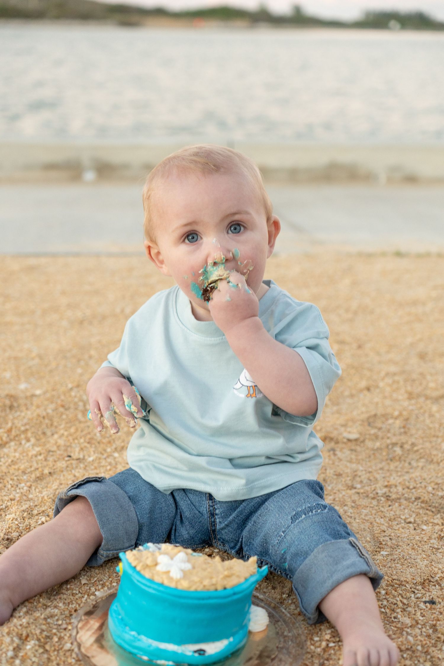 Baby sitting on beach sand eating smash cake, wearing light blue shirt and jeans, with ocean in background.