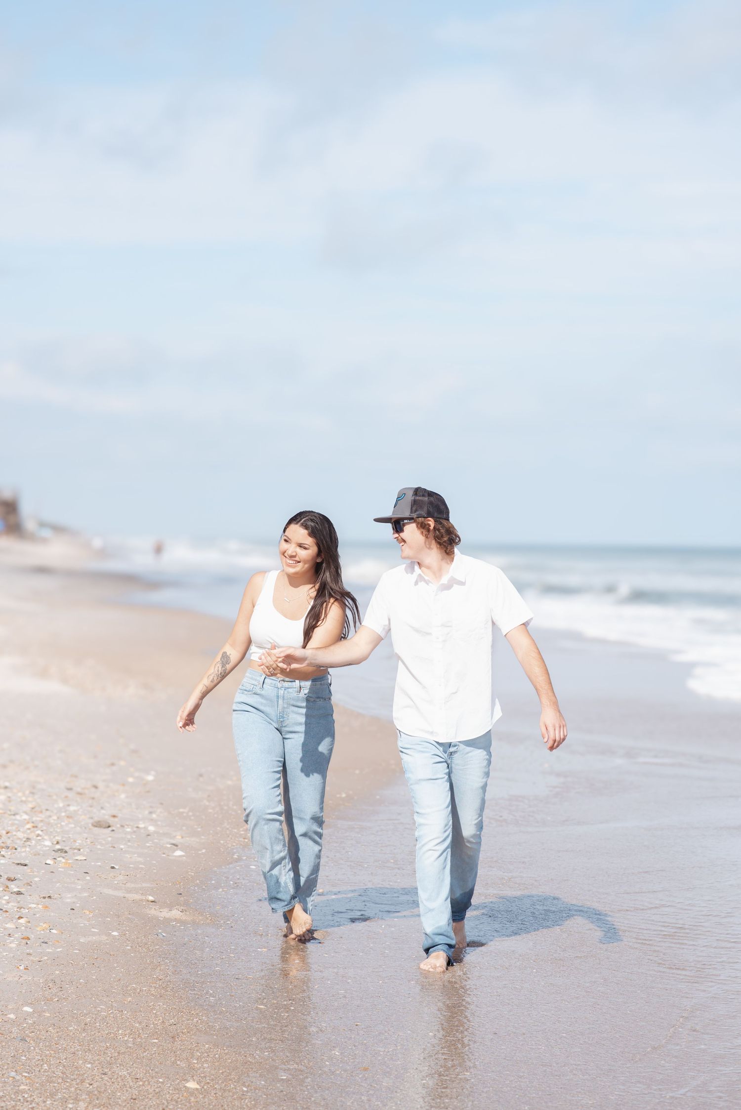 Couple walking hand in hand barefoot on a sunny beach, wearing jeans and white tops, smiling at each other.