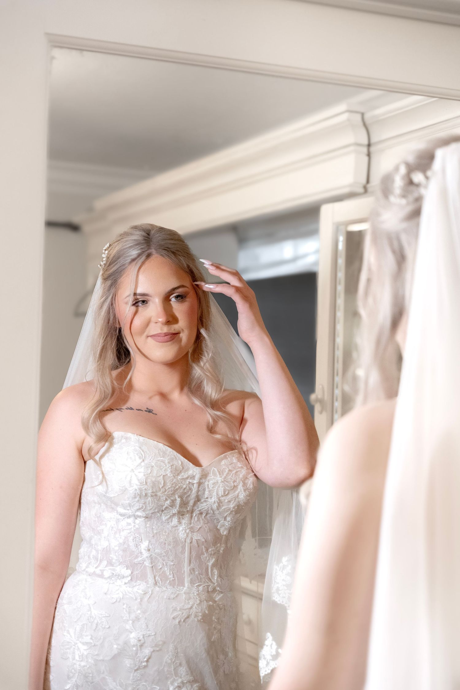 Bride in strapless lace wedding gown and veil adjusting hair while gazing into an elegant mirror.
