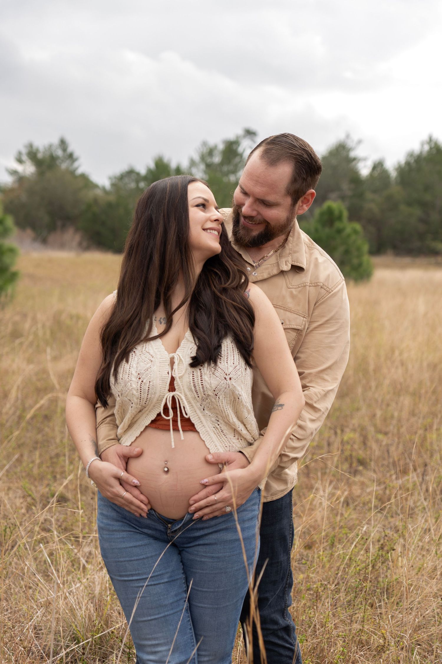 A smiling couple poses in a field, man embracing pregnant woman from behind, both looking at each other lovingly.