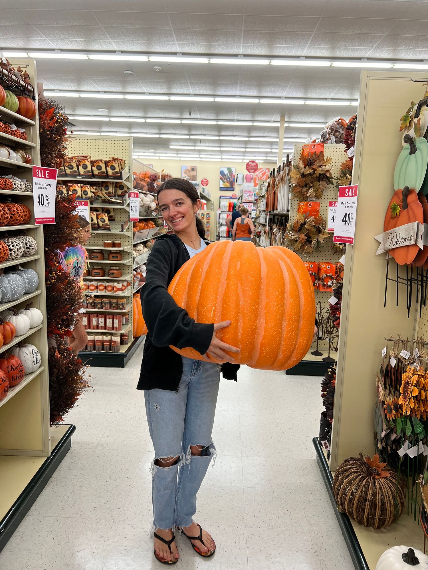 A smiling girl holds a large orange decorative pumpkin in a festive store aisle.