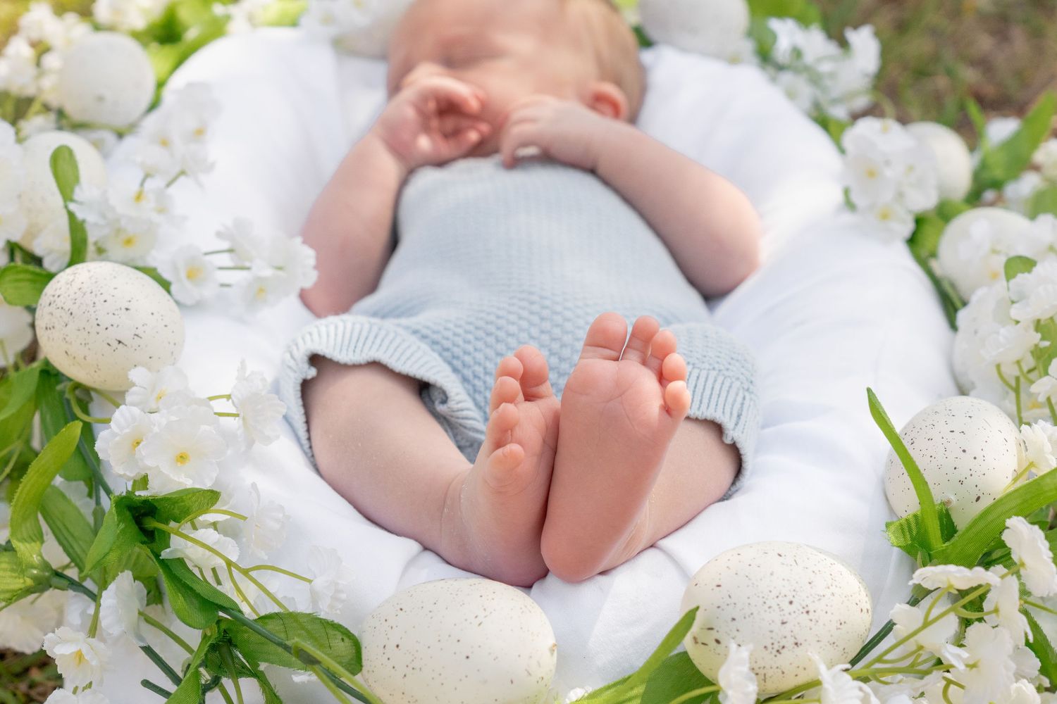 Newborn baby sleeping peacefully in a white nest surrounded by white flowers and Easter eggs outdoors.