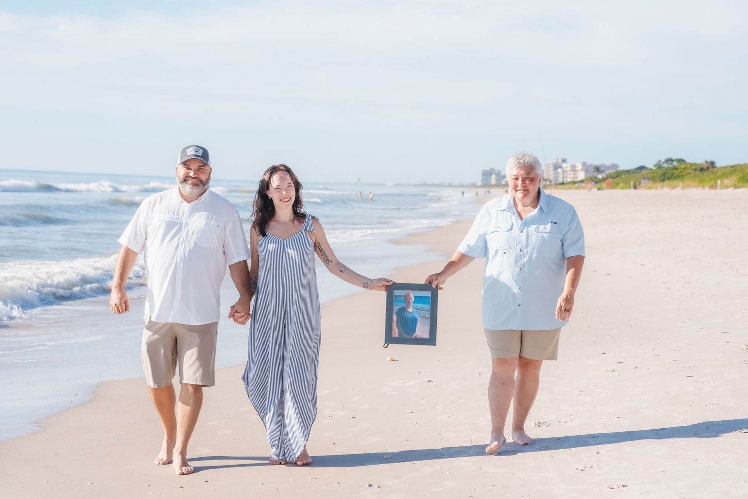 Three people walk on a sunny beach holding hands and a framed photo, waves and blue sky in the background.
