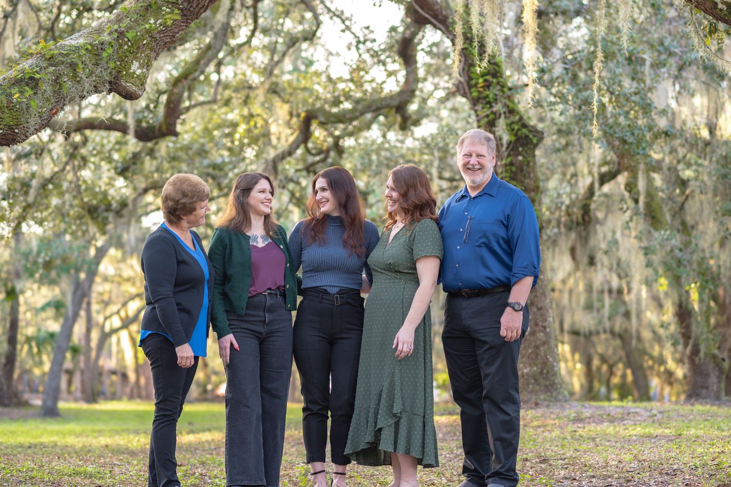 Family of five smiling outdoors under moss-draped oak trees in a lush green park setting.