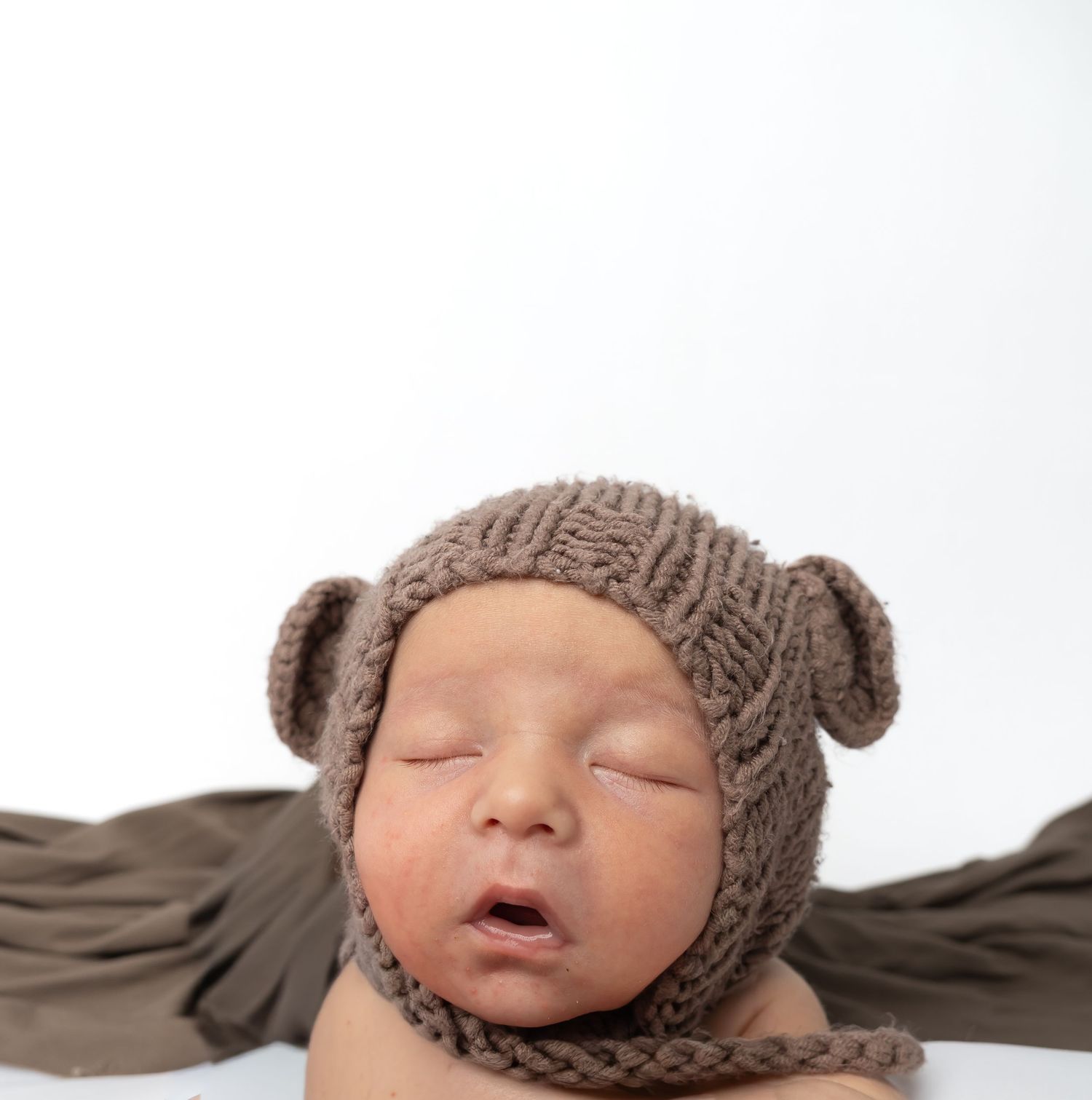 Newborn baby sleeping peacefully wearing a brown knitted bear ear bonnet hat on a white background.