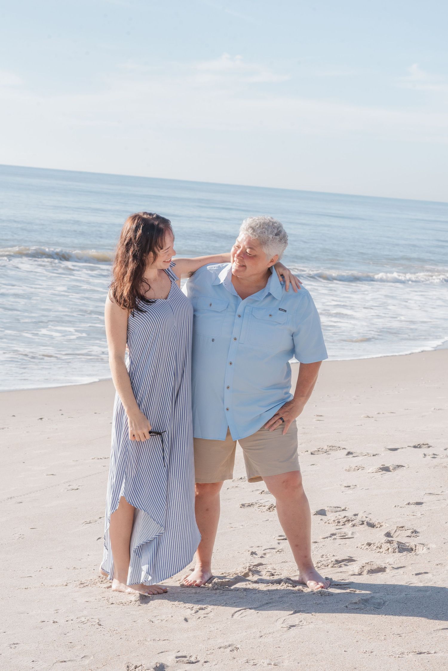 A couple stands barefoot on a sunny beach, smiling at each other, dressed in casual summer attire.