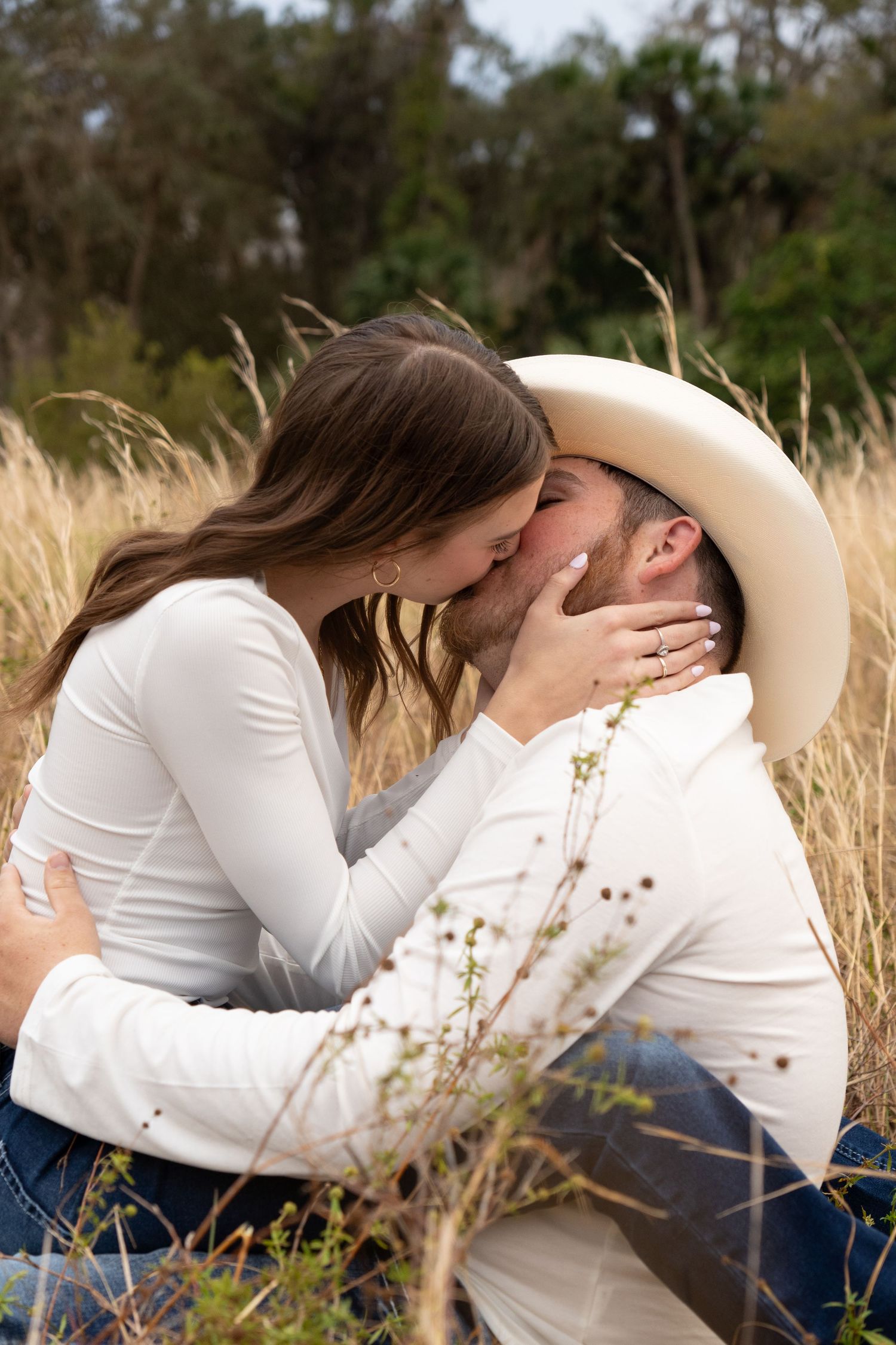 Couple sharing a romantic kiss in a golden wheat field, woman wearing white top and engagement ring, man in cowboy hat.