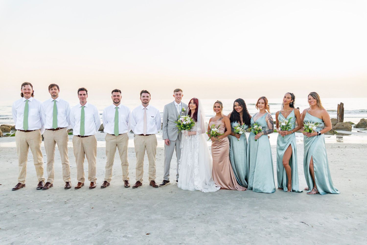Wedding party poses on beach at sunset, groomsmen in khaki with green ties, bridesmaids in sage and champagne gowns.