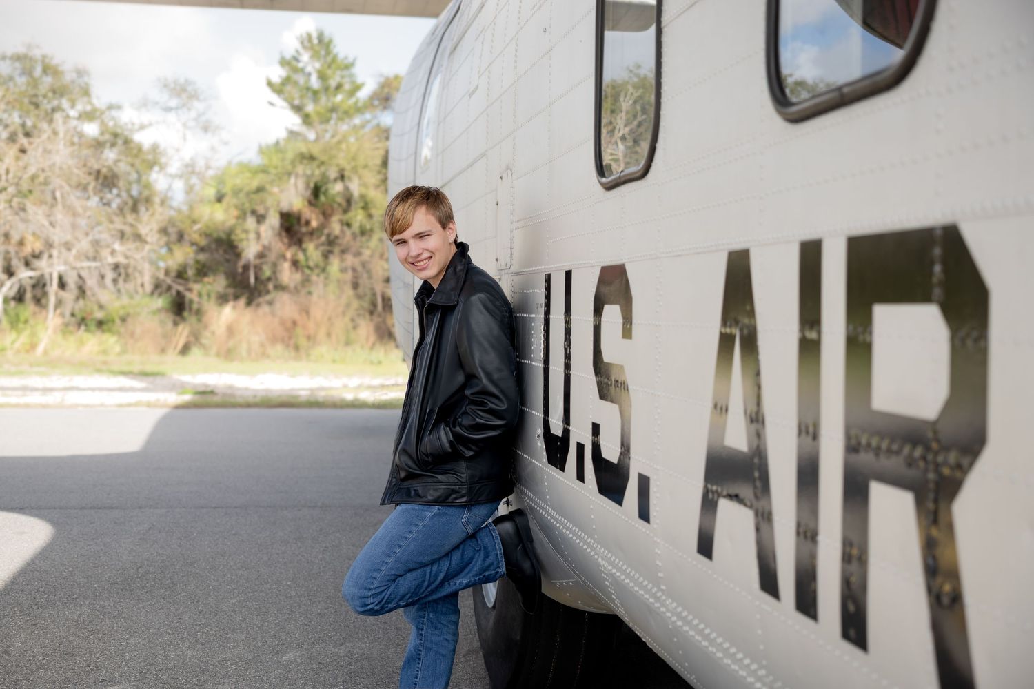 Teen boy in black jacket leaning against a US Air Force aircraft outdoors.