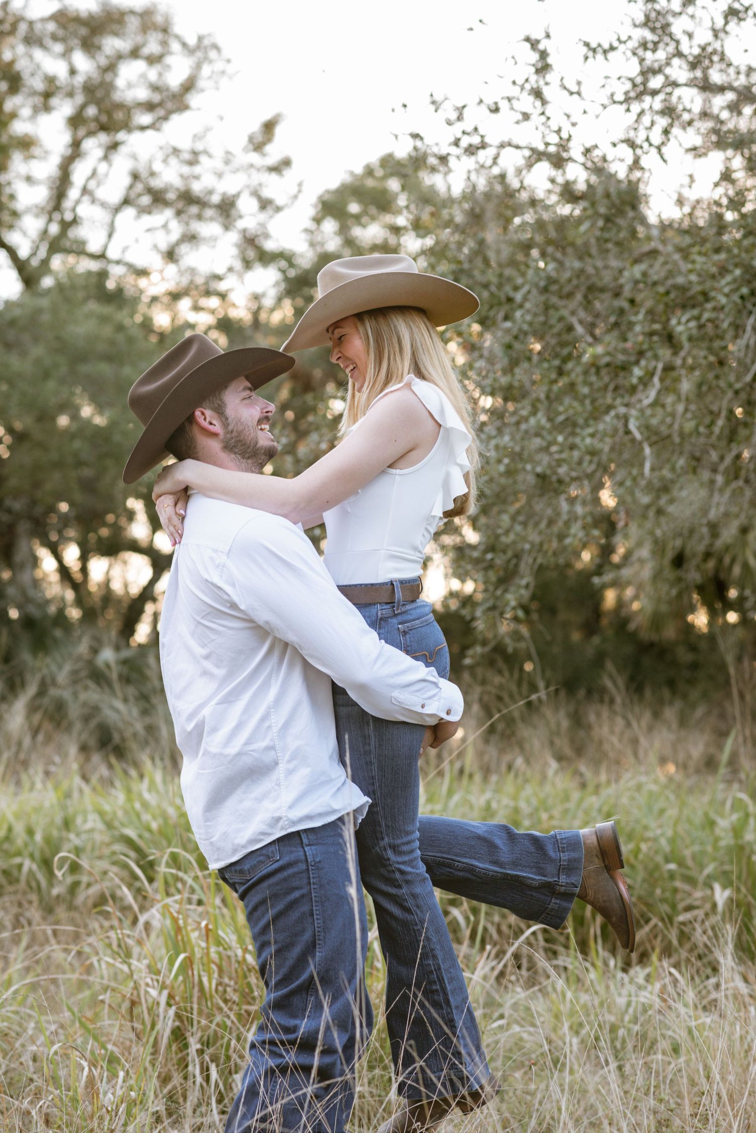 A couple in cowboy hats and white shirts embrace in a golden field, the man lifting the woman as they smile at each other.